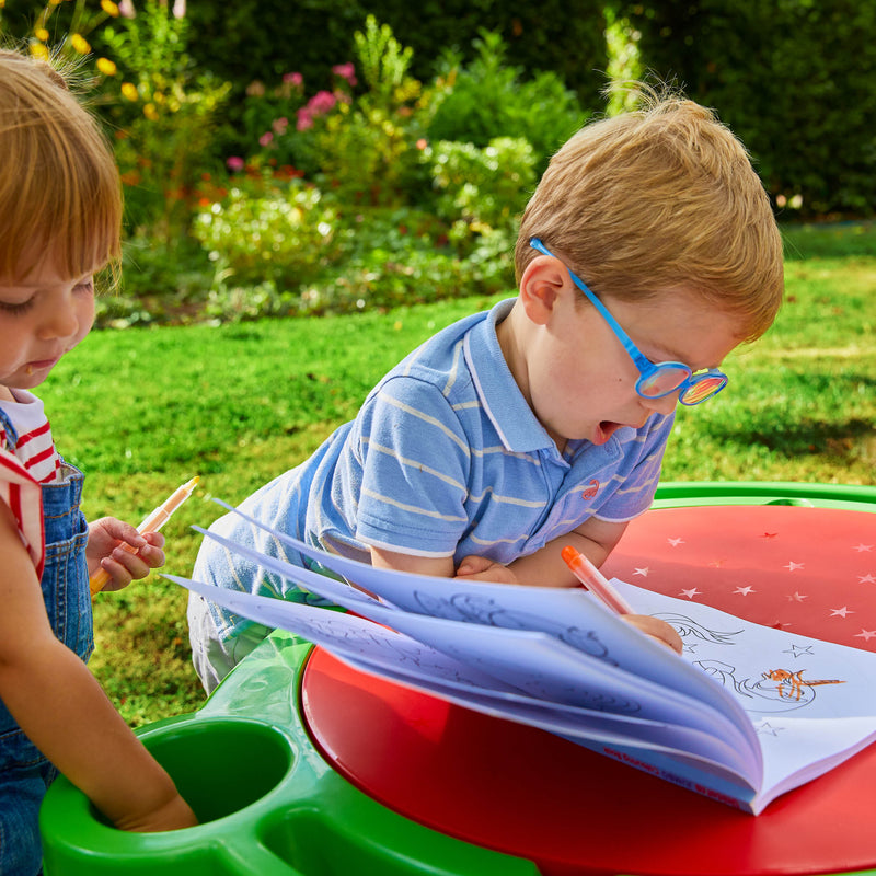 Des enfants dessinent sur un livre posé sur la table d’activités en plastique.