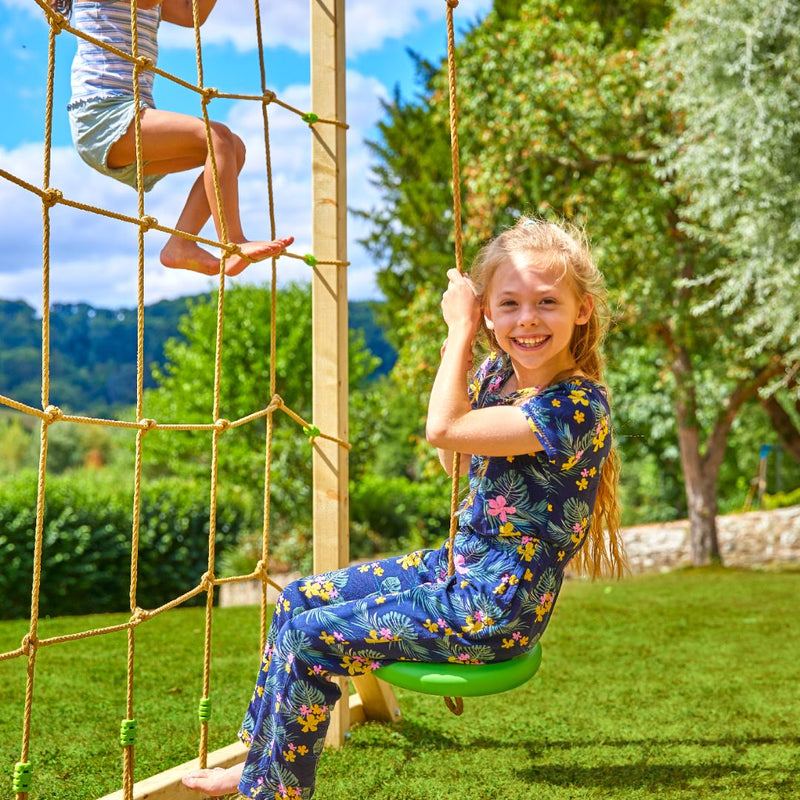 Deux enfants jouent sur une structure de jeux dans un jardin : une fillette est assise sur une tyrolienne, tandis qu’une autre enfant grimpe sur le filet.