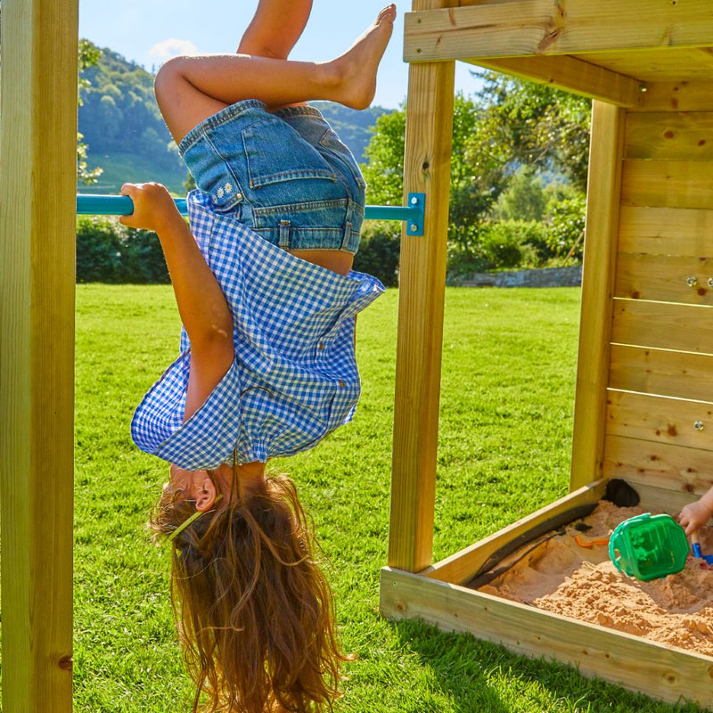 Une enfant se suspend la tête en bas à une barre de jeu sur une structure en bois, près d’un bac à sable en extérieur.