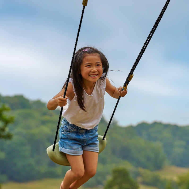 Enfant souriant sur une balançoire en plein air, jouant dans un jardin avec paysage en arrière-plan.