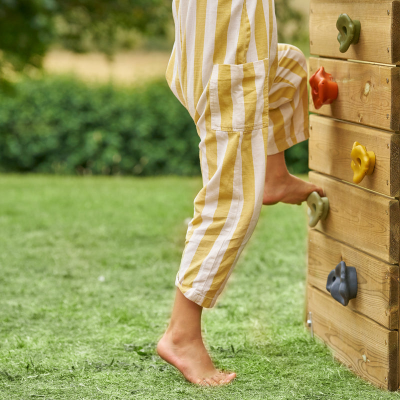 Enfant grimpant sur un mur d’escalade en bois avec prises colorées, intégré à une aire de jeux extérieure.