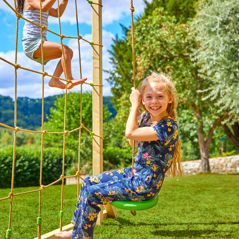 Deux enfants jouent sur une structure de jeux dans un jardin : une fillette est assise sur une tyrolienne, tandis qu’une autre enfant grimpe sur le filet.
