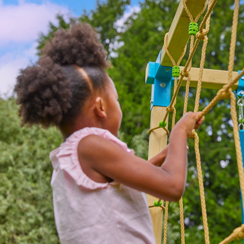 Enfant en train de grimper sur un filet fixé à une structure de jeux en bois, en extérieur.