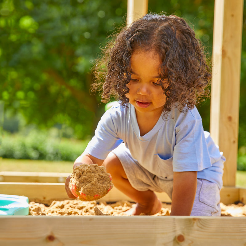 Un enfant joue dans un bac à sable en extérieur, tenant du sable dans ses mains.