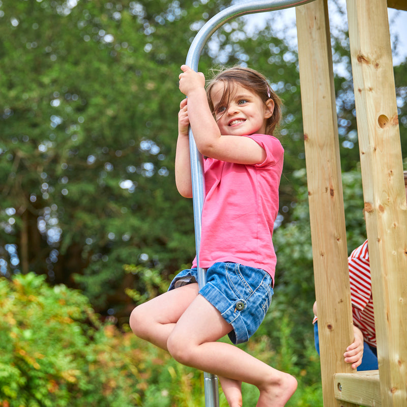 Une enfant descend la barre de pompier d’une aire de jeux en bois installée dans un jardin.