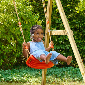 Une enfant se balance sur une balançoire rouge suspendue par des cordes, dans un jardin.