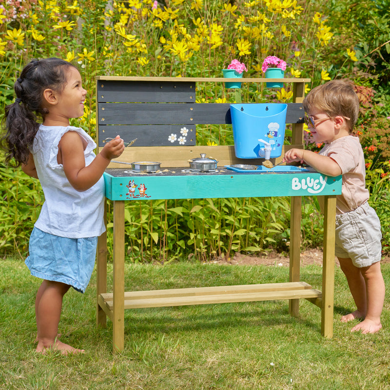 Deux enfants jouant ensemble avec une cuisine d’extérieur en bois décorée de personnages Bluey, équipée de plaques de cuisson factices, casseroles, évier avec réservoir d’eau et étagère, installée dans un jardin fleuri.
