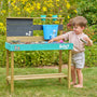 Enfant jouant avec une cuisine d’extérieur en bois décorée de personnages Bluey, avec plan de travail turquoise, évier et accessoires, installée dans un jardin avec pelouse et fleurs en arrière-plan.