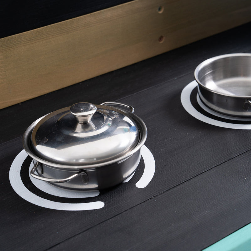 Détail du plan de travail d’une cuisine d’extérieur pour enfant, avec deux plaques de cuisson factices peintes et des casseroles en métal, vu de près.