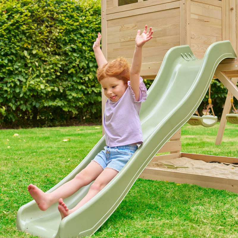 Enfant glissant les bras levés sur un toboggan vert kaki, installé dans un jardin avec pelouse et structure de jeu en bois à l’arrière-plan.