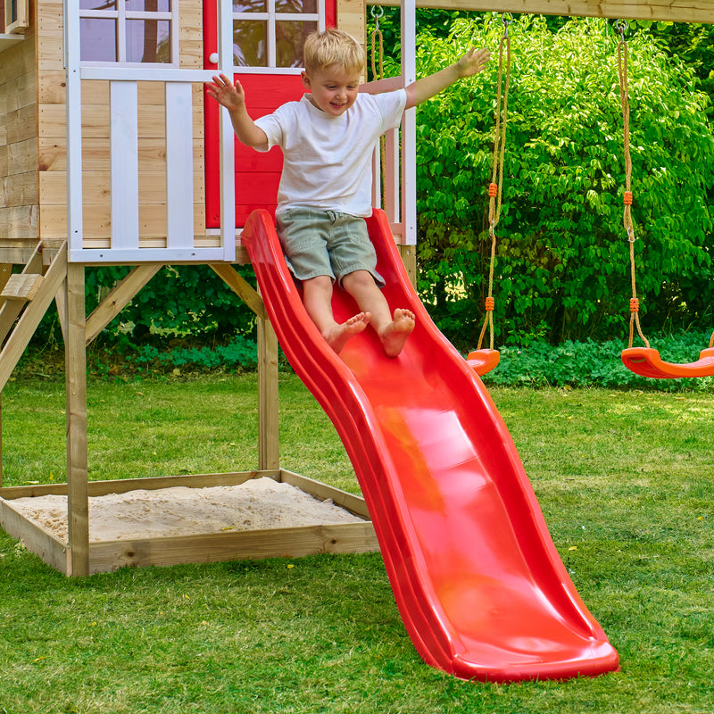 Enfant descendant un toboggan en plastique rouge fixé à une maisonnette en bois, installé dans un jardin avec pelouse, bac à sable et balançoires à l’arrière-plan.