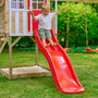 Enfant descendant un toboggan en plastique rouge fixé à une maisonnette en bois, installé dans un jardin avec pelouse, bac à sable et balançoires à l’arrière-plan.