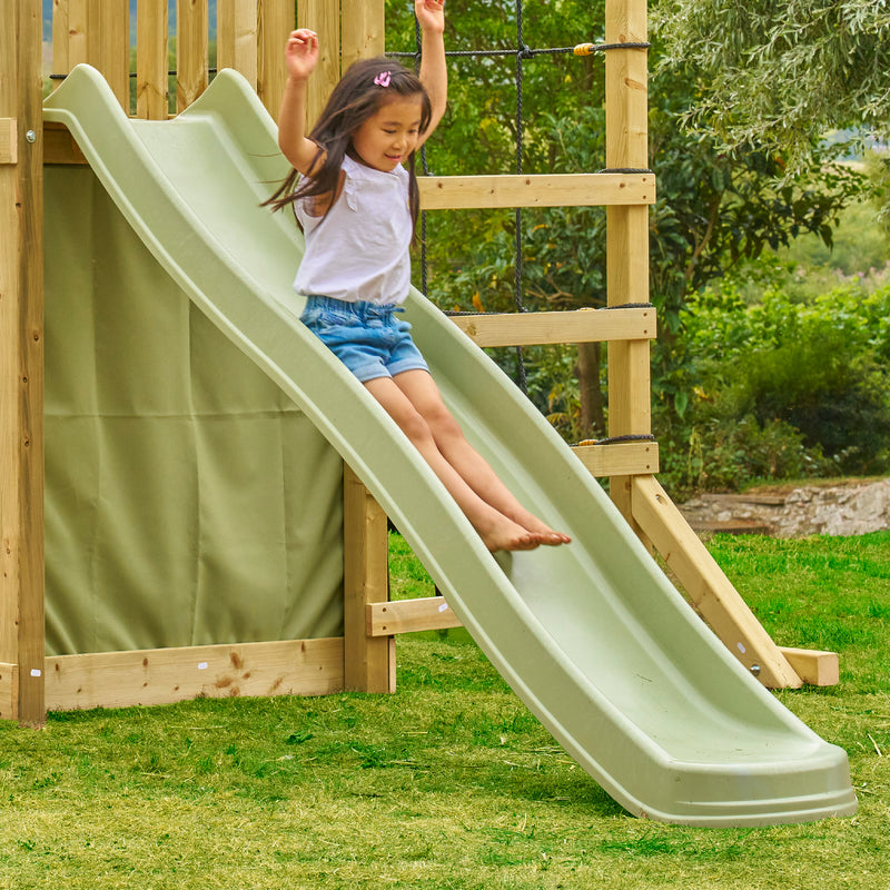 Enfant descendant un toboggan en plastique vert kaki fixé à une structure de jeu en bois, installé dans un jardin avec pelouse et arbres en arrière-plan.