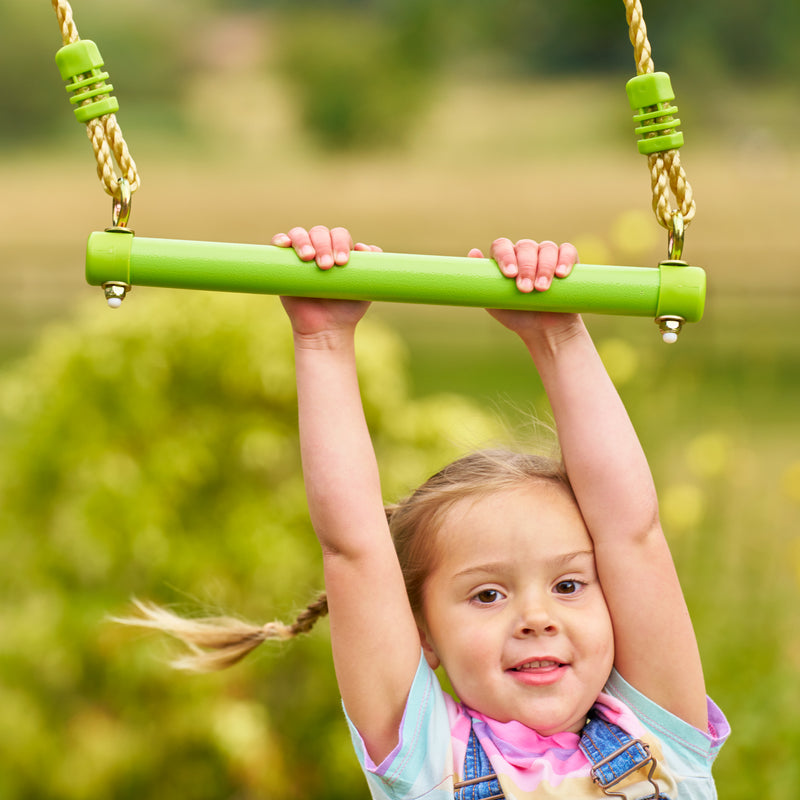 Une enfant se suspend au trapèze en métal TP Toys, conçu pour développer coordination, force et équilibre.
