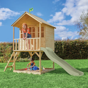 Une enfant est en haut d’une cabane en bois sur pilotis, une autre glisse sur le toboggan et un enfant joue dans le bac à sable situé en dessous de la cabane.