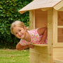 Une enfant passe la tête par la fenêtre d’une maisonnette en bois installée dans un jardin.