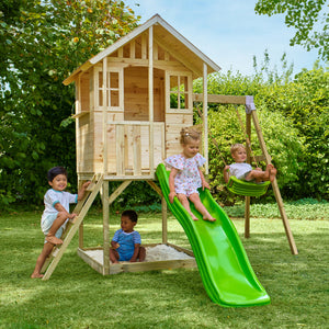 Quatre enfants jouent dans un jardin avec une maisonnette en bois sur pilotis, équipée d’une échelle, d’un bac à sable, d’un toboggan vert et d’une balançoire.
