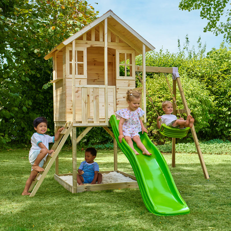 Quatre enfants jouent dans un jardin avec une maisonnette en bois sur pilotis, équipée d’une échelle, d’un bac à sable, d’un toboggan vert et d’une balançoire.