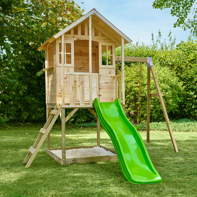 Maisonnette en bois sur pilotis dans un jardin, équipée d’une échelle, d’un bac à sable, d’un toboggan vert et d’une balançoire.