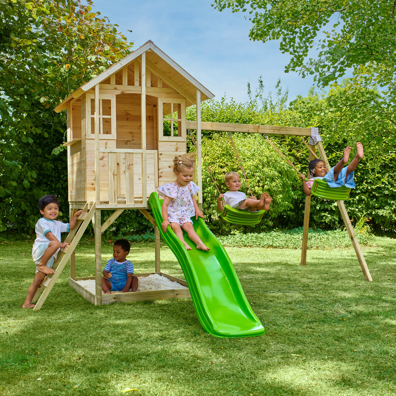 Cinq enfants jouent dans un jardin avec une maisonnette en bois sur pilotis, équipée d’une échelle, d’un bac à sable, d’un toboggan vert et de deux balançoires.