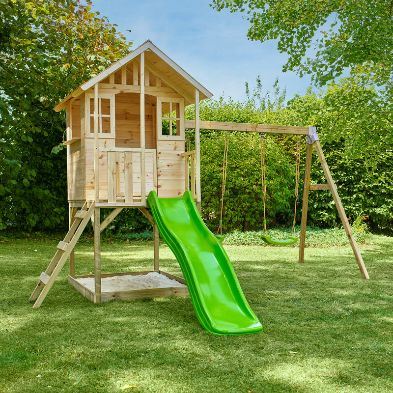 Maisonnette en bois sur pilotis dans un jardin, équipée d’une échelle, d’un bac à sable, d’un toboggan vert et dd deux balançoires.