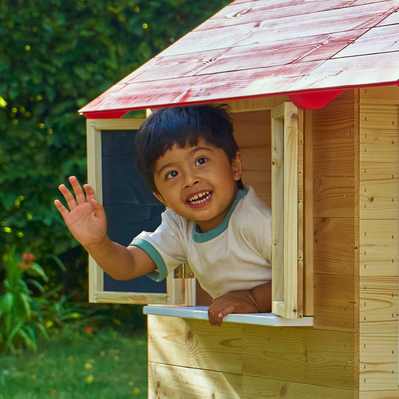 Enfant faisant signe par la fenêtre ouverte d’une maisonnette en bois avec volets, dans un jardin.
