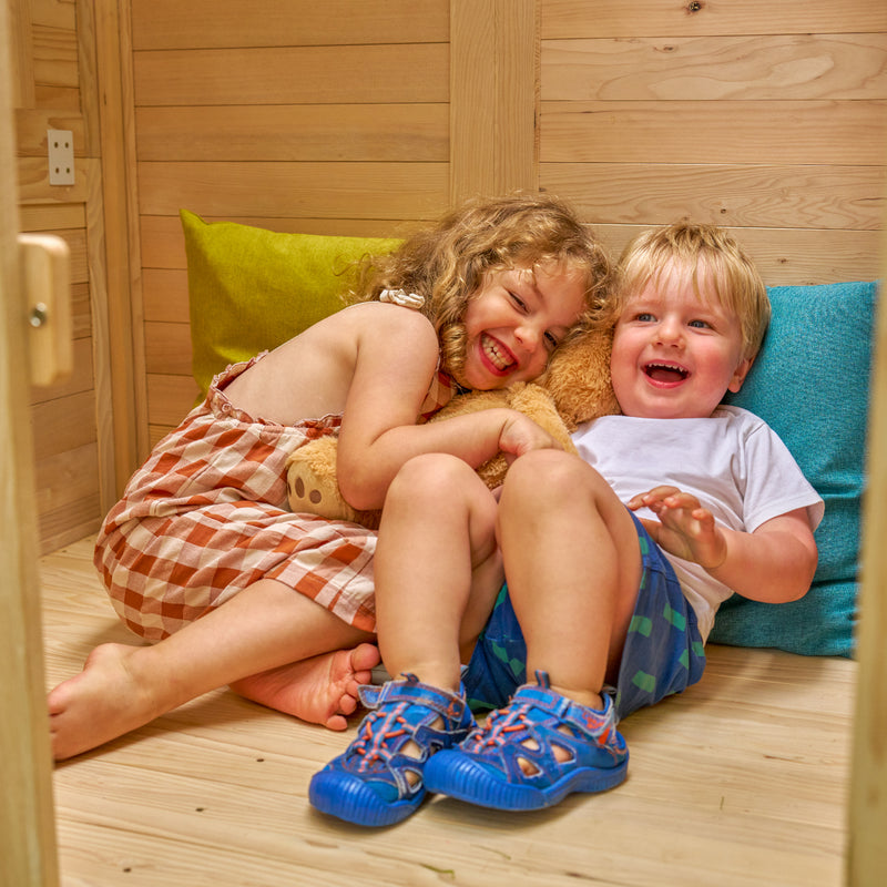 Deux enfants assis à l’intérieur de la maisonnette rient en jouant.