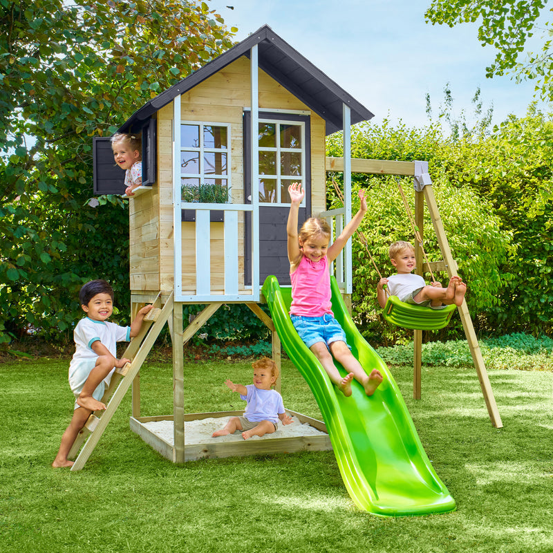 Des enfants jouent autour d’une maisonnette en bois sur pilotis avec façade grise et blanche, équipée d’une échelle, d’un bac à sable d'une balançoire et d’un toboggan vert.