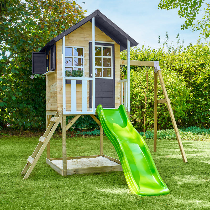 Maisonnette en bois sur pilotis avec façade grise et blanche, équipée d’une échelle, d’un bac à sable, d’un toboggan vert et d'une balançoire dans un jardin.