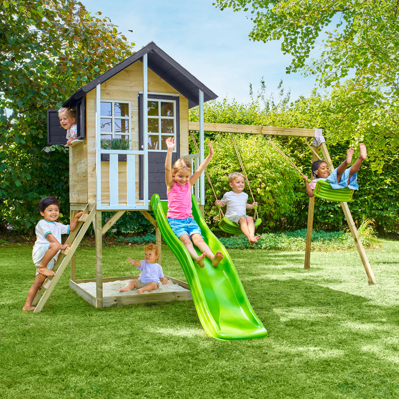 Des enfants jouent autour d’une maisonnette en bois sur pilotis avec façade grise et blanche, équipée d’une échelle, d’un bac à sable, de deux balançoires et d’un toboggan vert.