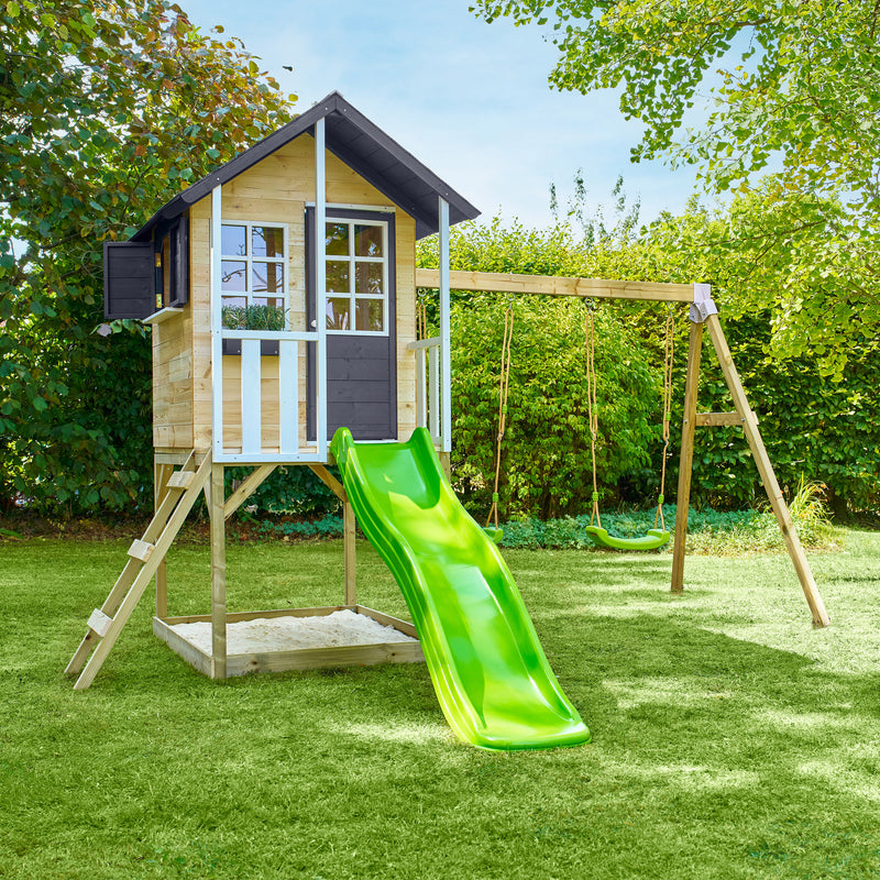 Maisonnette en bois sur pilotis avec façade grise et blanche, équipée d’une échelle, d’un bac à sable, d’un toboggan vert et de deux balançoires dans un jardin.