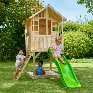 Trois enfants jouent dans un jardin avec une maisonnette en bois sur pilotis, équipée d’une échelle, d’un bac à sable et d’un toboggan vert.