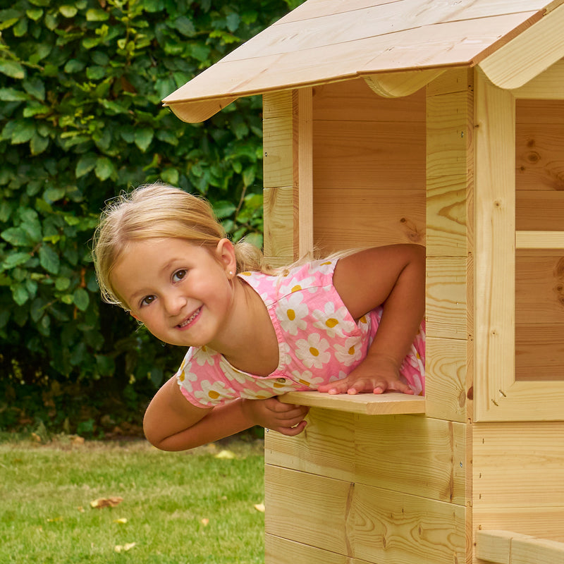 Une enfant passe la tête par la fenêtre d’une maisonnette en bois installée dans un jardin.
