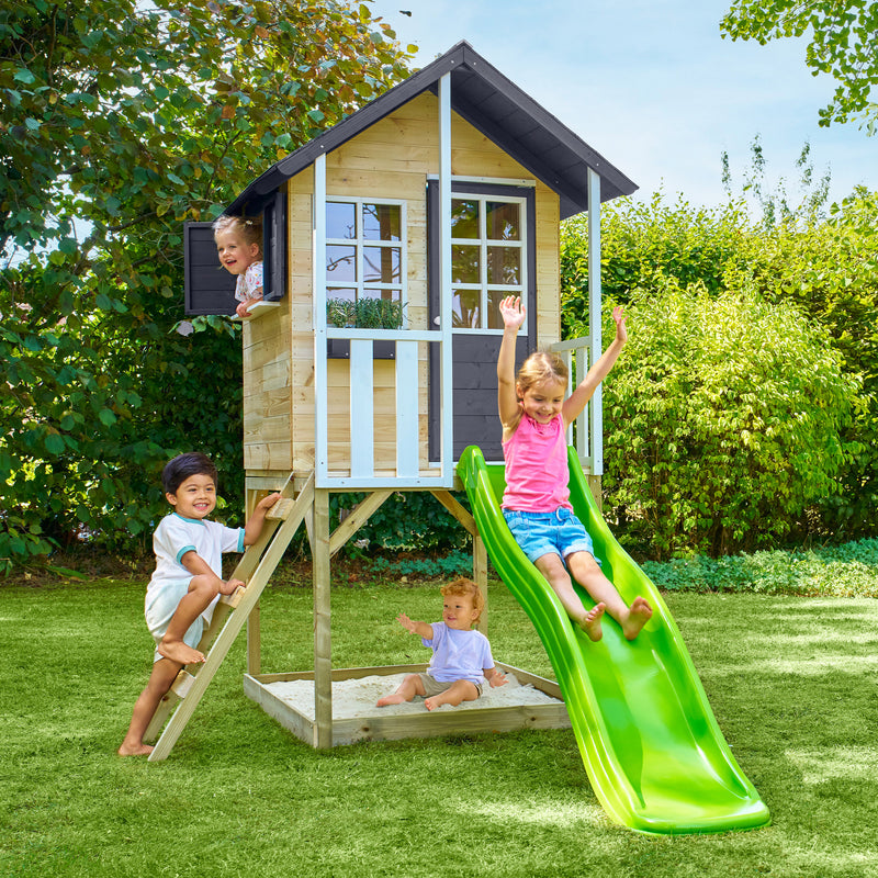 Des enfants jouent autour d’une maisonnette en bois sur pilotis, équipée d’une échelle, d’un bac à sable et d’un toboggan vert.
