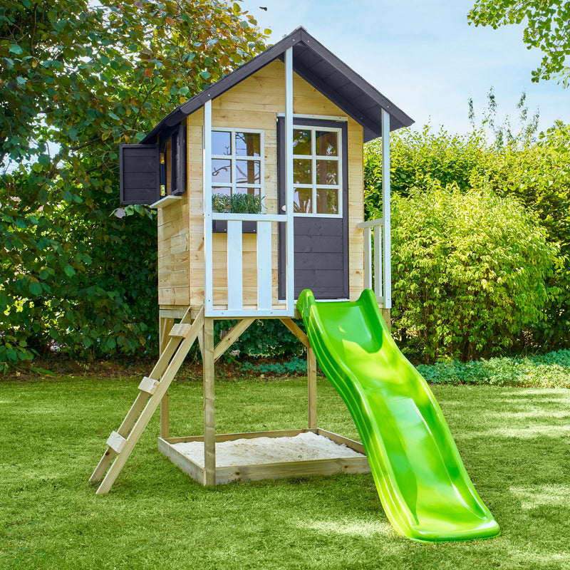 Maisonnette en bois sur pilotis, équipée d’une échelle, d’un bac à sable et d’un toboggan vert dans un jardin.