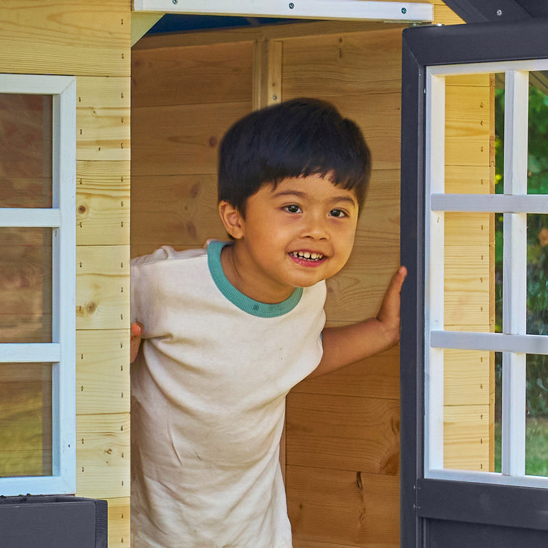 Enfant souriant debout dans l’entrée d’une maisonnette en bois, avec porte ouverte.