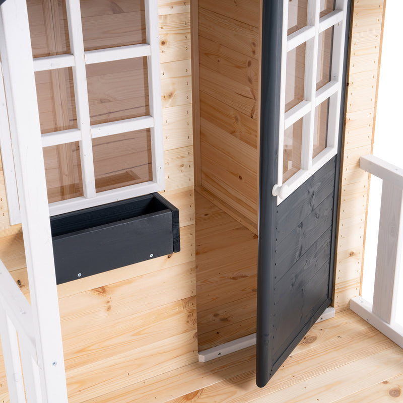 Détail de l’entrée d’une maisonnette en bois avec porte gris foncé entrouverte, fenêtre blanche et petit bac à l'extérieur.