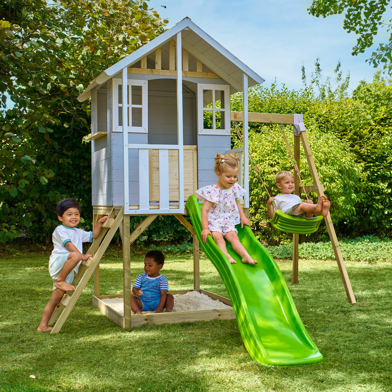 Des enfants jouent autour d’une maisonnette en bois sur pilotis avec façade grise et blanche, équipée d’une échelle, d’un bac à sable d'une balançoire et d’un toboggan vert.
