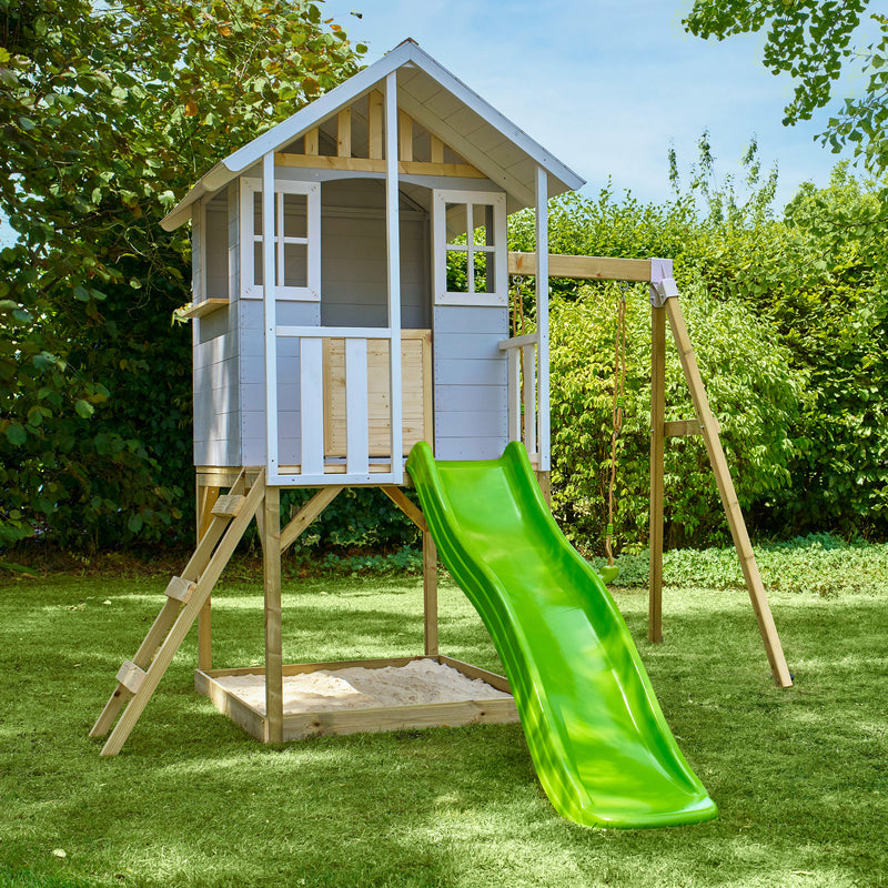Maisonnette en bois sur pilotis avec façade grise et blanche, équipée d’une échelle, d’un bac à sable, d’un toboggan vert et d'une balançoire dans un jardin.