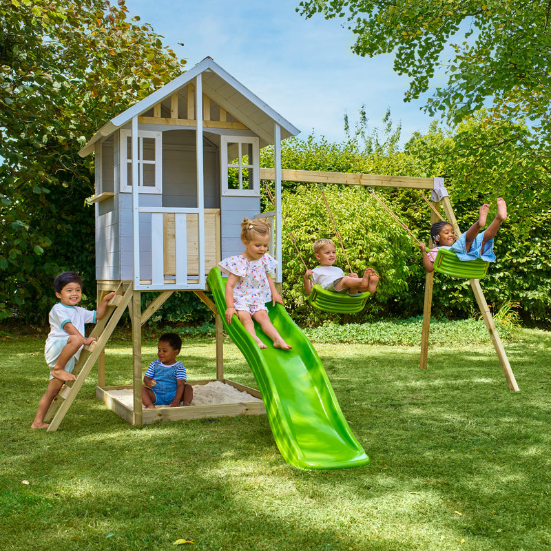 Des enfants jouent autour d’une maisonnette en bois sur pilotis avec façade grise et blanche, équipée d’une échelle, d’un bac à sable, de deux balançoires et d’un toboggan vert.