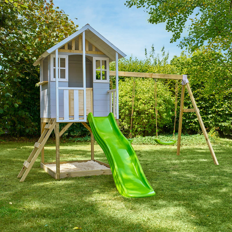Maisonnette en bois sur pilotis avec façade grise et blanche, équipée d’une échelle, d’un bac à sable, d’un toboggan vert et de deux balançoires dans un jardin.
