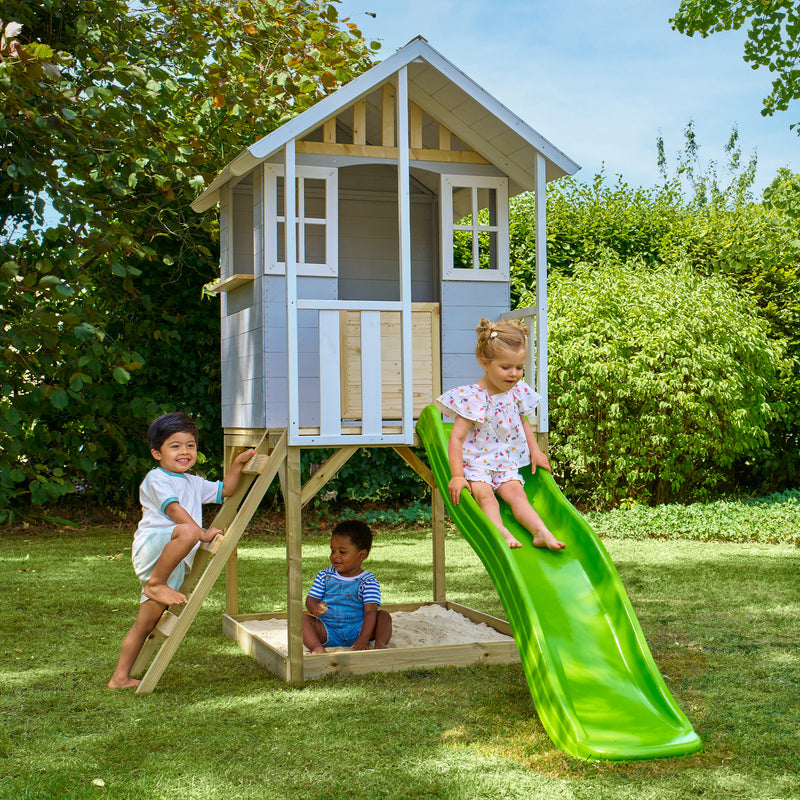 Des enfants jouent autour d’une maisonnette en bois sur pilotis avec façade grise et blanche, équipée d’une échelle, d’un bac à sable et d’un toboggan vert.