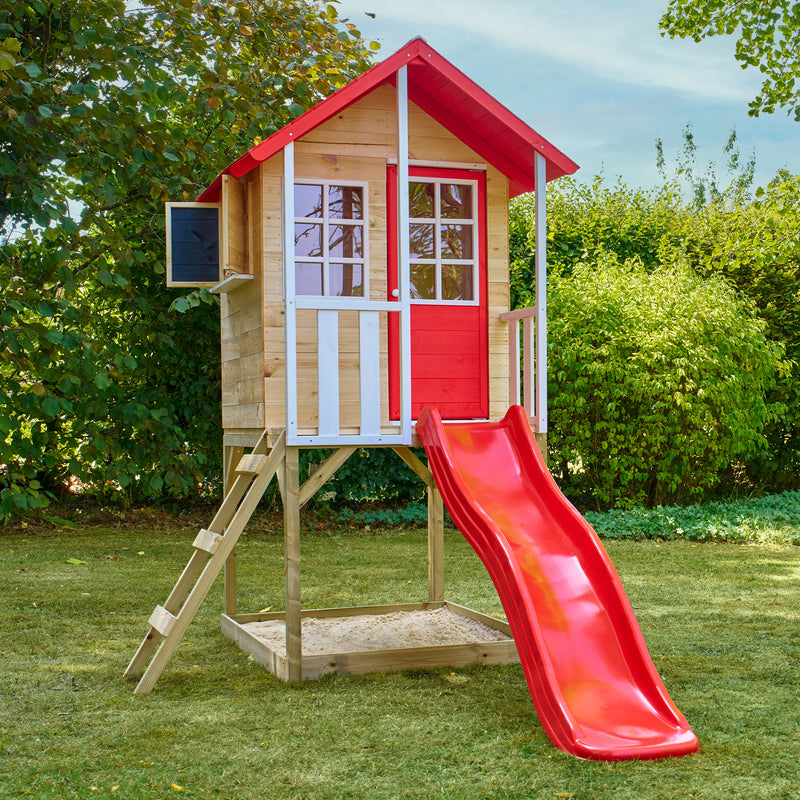 Maisonnette en bois sur pilotis, équipée d’une échelle, d’un bac à sable et d’un toboggan rouge dans un jardin.