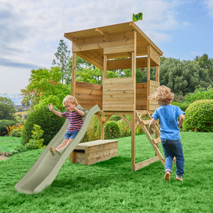 Maisonnette cabane sur pilotis en bois avec toboggan dans un jardin : un enfant glisse sur le toboggan tandis qu’un autre court vers l’échelle de la maisonnette.