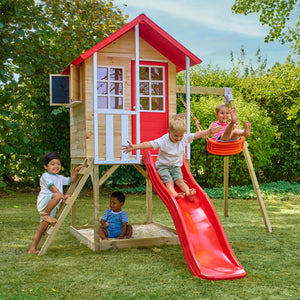 Des enfants jouent autour d’une maisonnette en bois sur pilotis avec façade rouge et blanche, équipée d’une échelle, d’un bac à sable d'une balançoire et d’un toboggan rouge.
