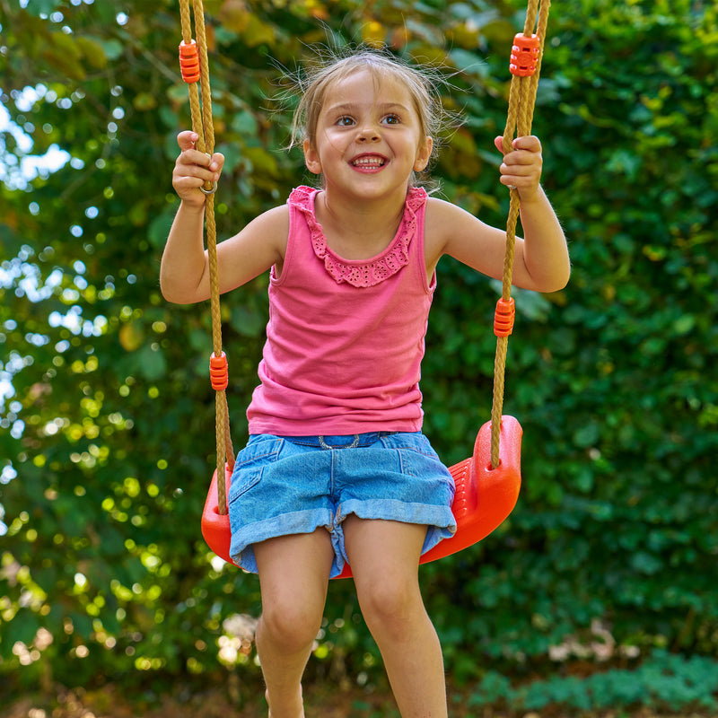 Une enfant se balance sur une balançoire rouge suspendue par des cordes, dans un jardin.