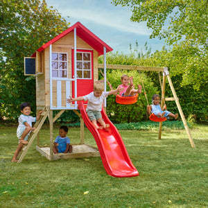 Des enfants jouent autour d’une maisonnette en bois sur pilotis avec façade rouge et blanche, équipée d’une échelle, d’un bac à sable, de deux balançoires et d’un toboggan rouge.