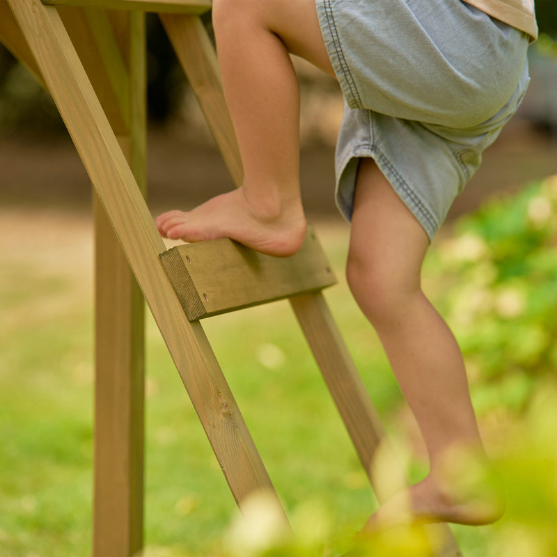 Gros plan sur les pieds d’un enfant montant l’échelle en bois d’une maisonnette sur pilotis dans un jardin.