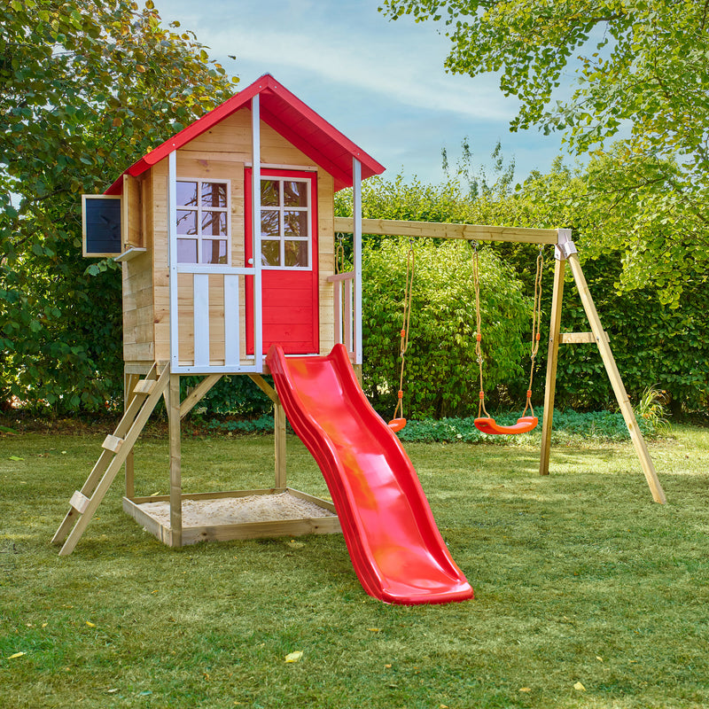 Maisonnette en bois sur pilotis avec façade grise et blanche, équipée d’une échelle, d’un bac à sable, d’un toboggan vert et de deux balançoires dans un jardin.