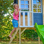Enfant montant l’échelle d’accès d’une maisonnette surélevée en bois avec rambarde blanche et toboggan vert.